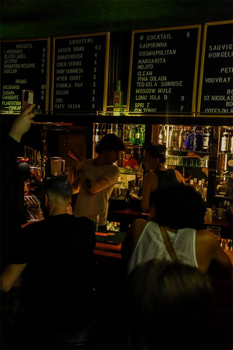 Un bar animé avec des clients et un barman, et un tableau noir listant des boissons.