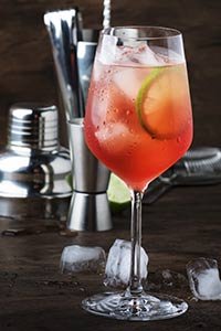 Un cocktail coloré avec des tranches de citron vert, servi dans un verre à vin, entouré de glaçons et d’accessoires de bar.