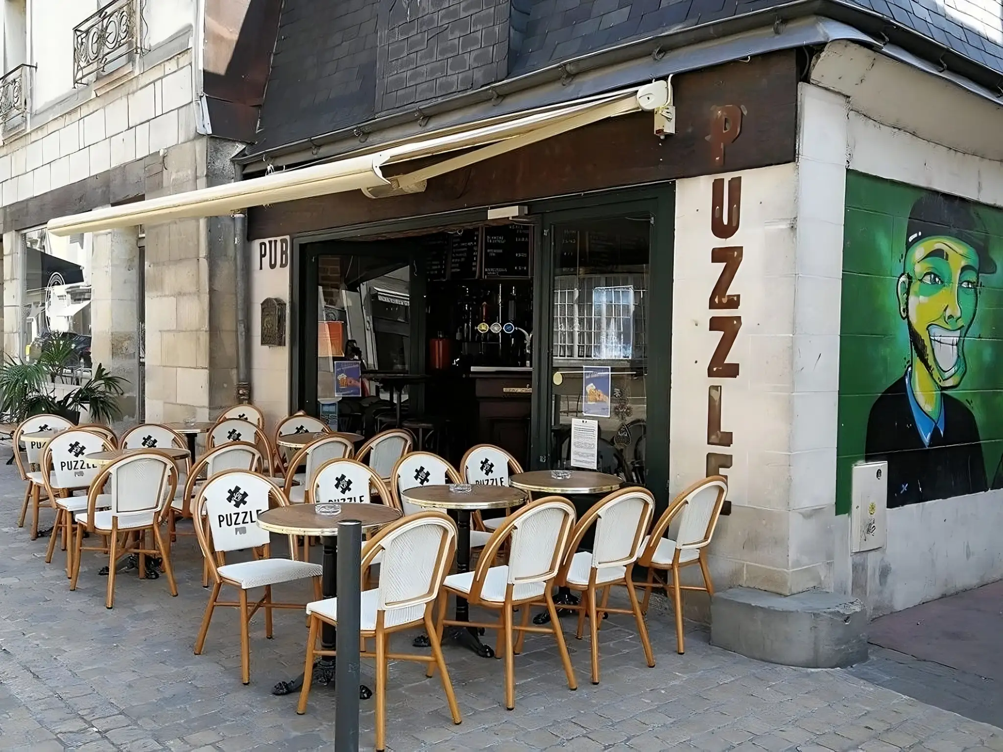 Une vue extérieure du Puzzle Pub à Tours avec des chaises en bois disposées devant et une peinture murale colorée.